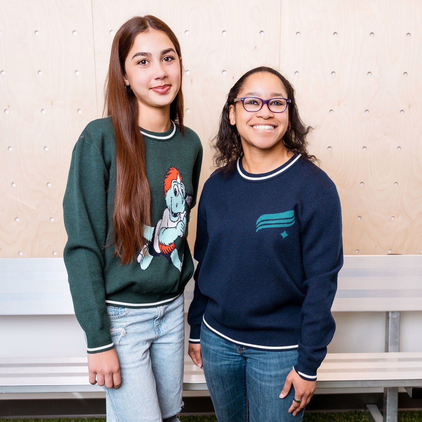 Two people standing next to each other in a casual setting with a bench and wall in the background.