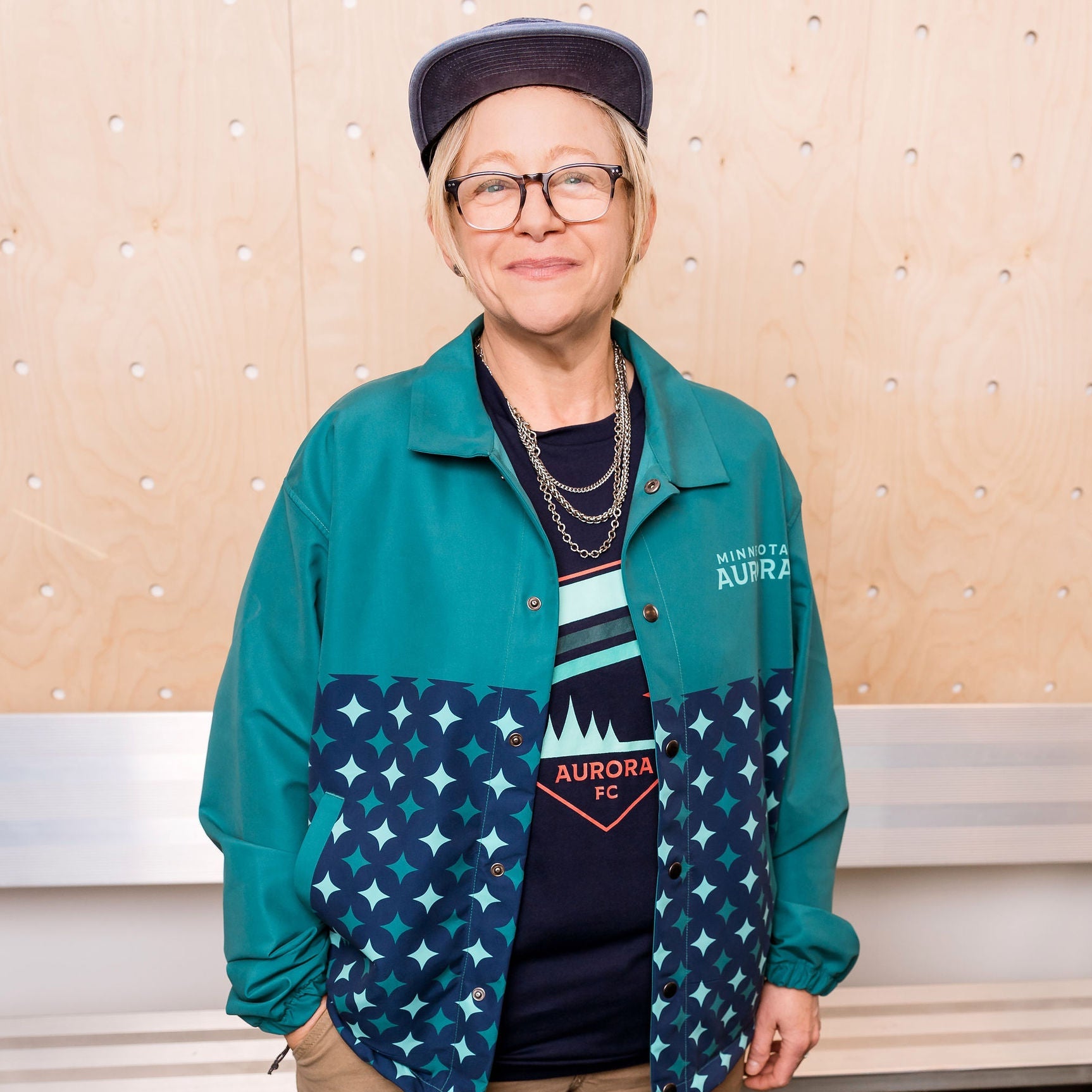 Person wearing a teal jacket with star pattern, beige pants, and a blue cap indoors.