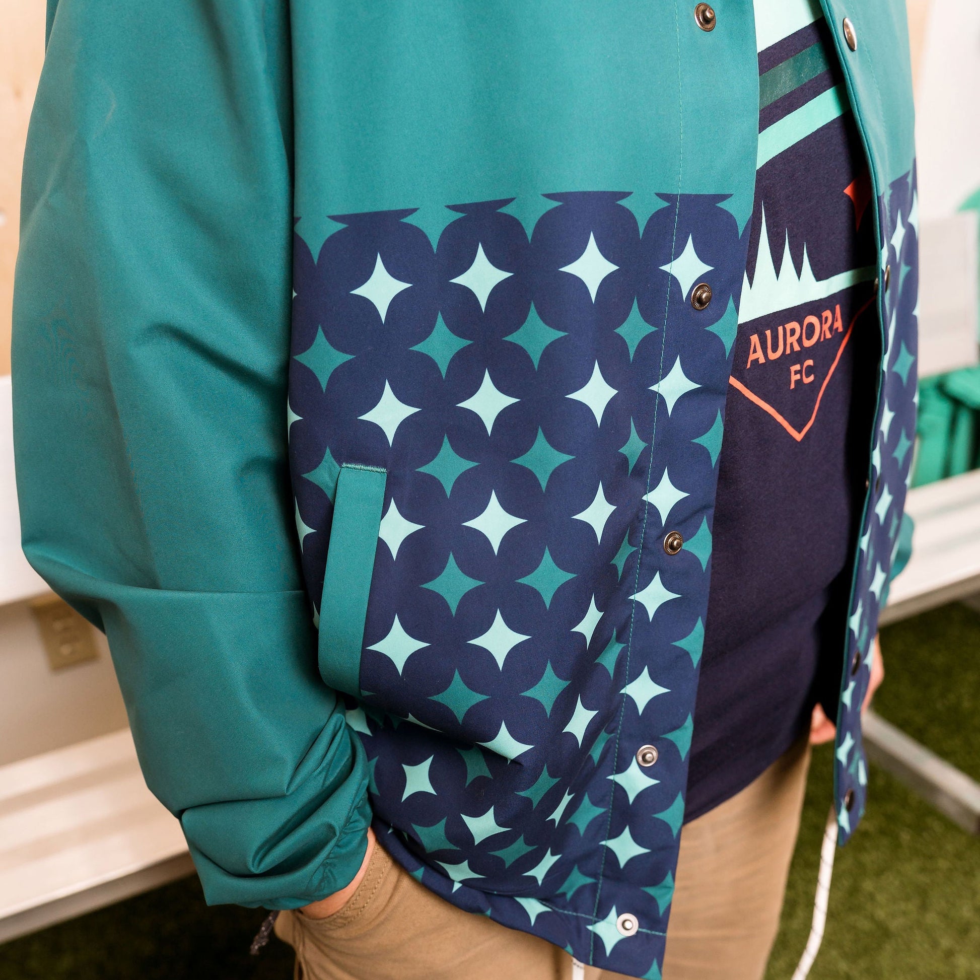 Person wearing a teal jacket with a patterned design over a dark shirt with 'Aurora FC' text.
