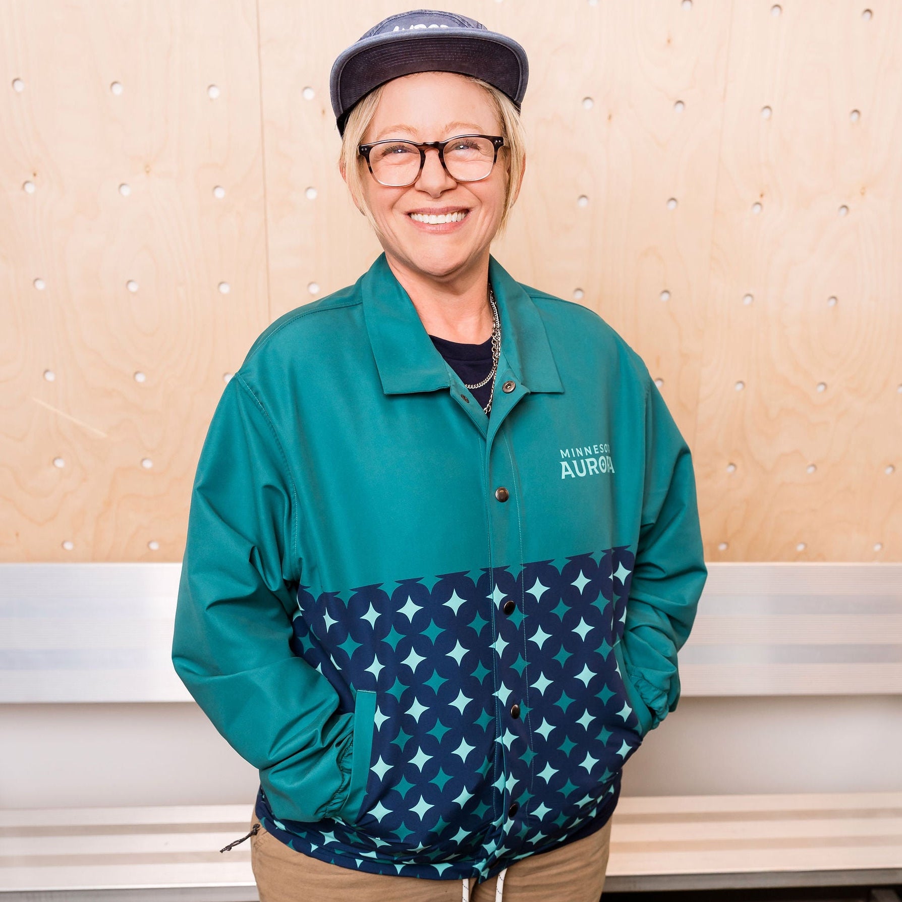 Person wearing a teal jacket with star pattern, beige pants, standing in front of a white wall.