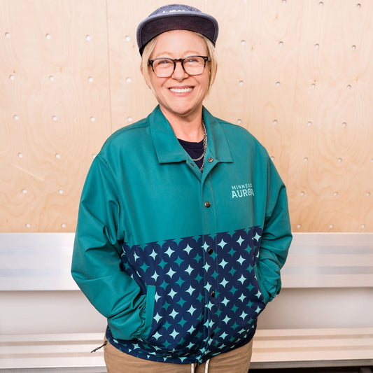 Person wearing a teal jacket with star pattern, beige pants, standing in front of a white wall.