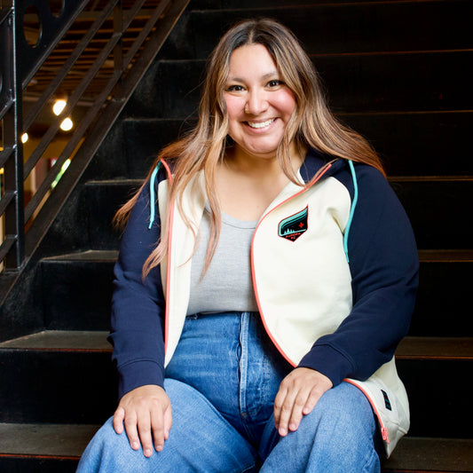 A woman smiling while sitting on dark wooden stairs, wearing a cream and navy zip-up vest over a gray top and blue jeans.