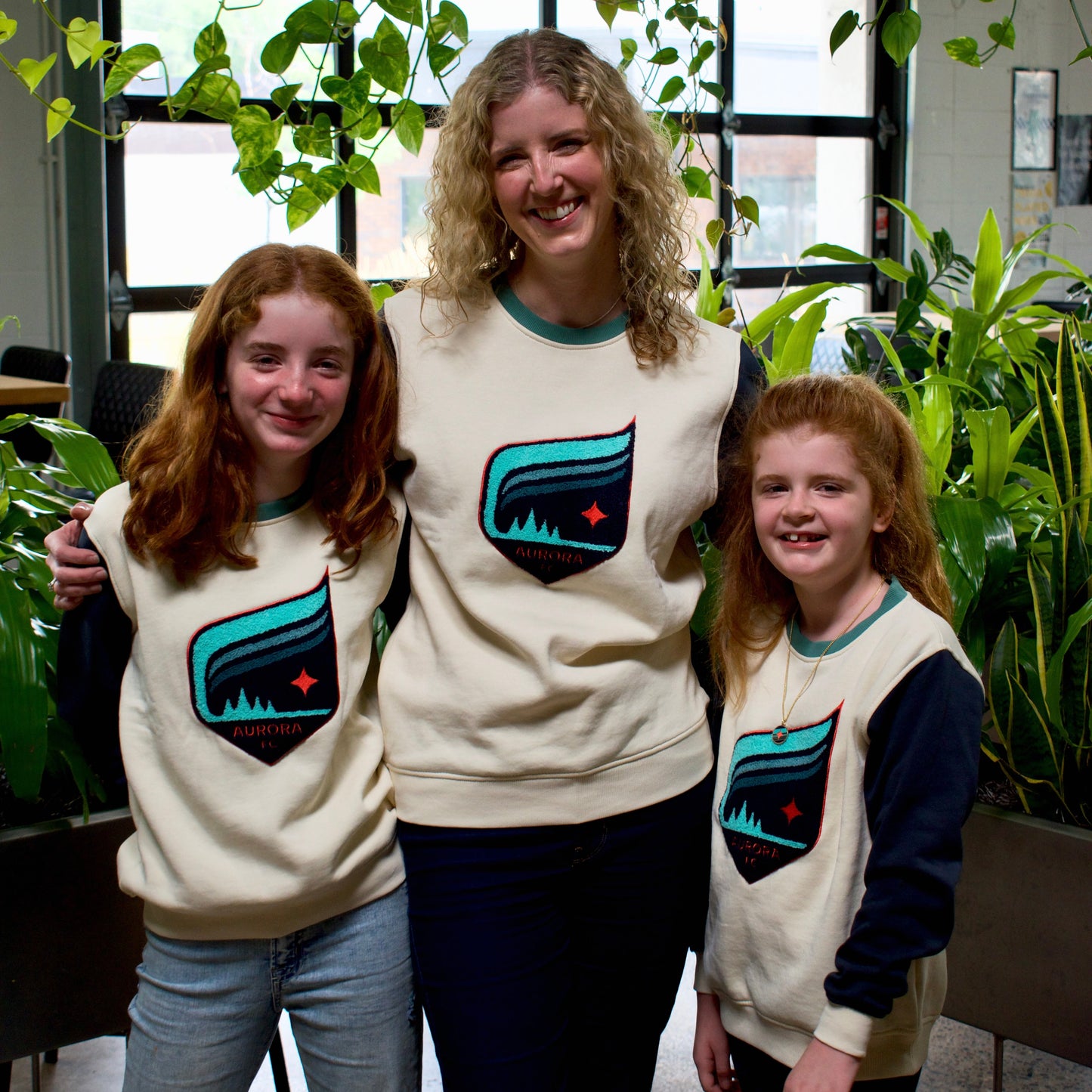 Three people posing together indoors wearing matching cream sweatshirts with a teal and black mountain graphic and a small red star logo. The setting features green plants and large windows in the background.