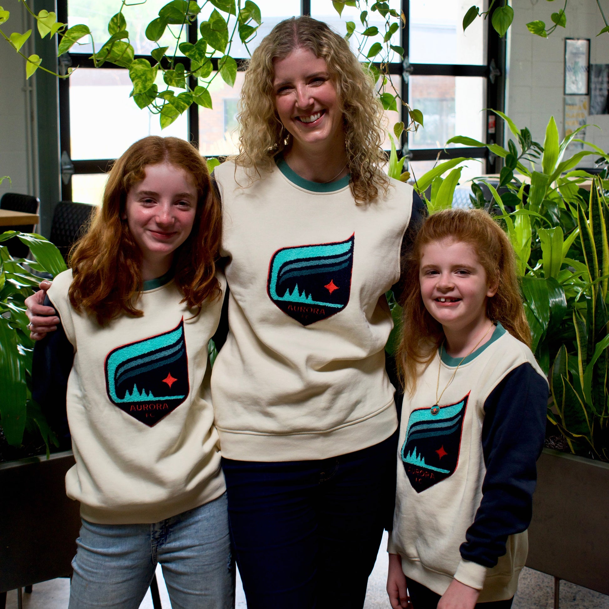 Three people posing together indoors wearing matching cream sweatshirts with a teal and black mountain graphic and a small red star logo. The setting features green plants and large windows in the background.