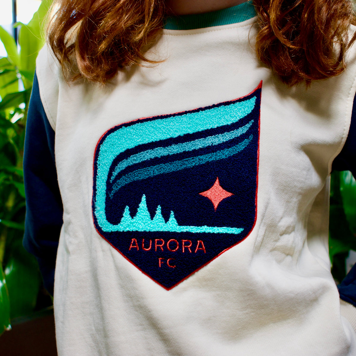 A person wearing a cream and navy long-sleeve sweatshirt with a large embroidered shield patch on the chest featuring a teal and navy aurora design and the text 'AURORA FC'.