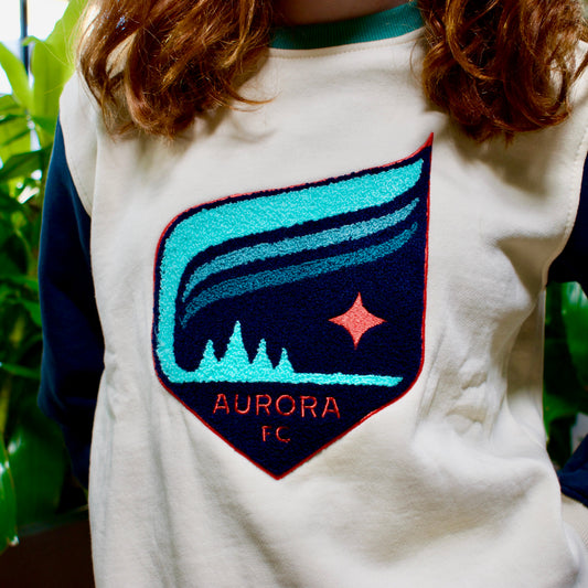 A person wearing a cream and navy long-sleeve sweatshirt with a large embroidered shield patch on the chest featuring a teal and navy aurora design and the text 'AURORA FC'.