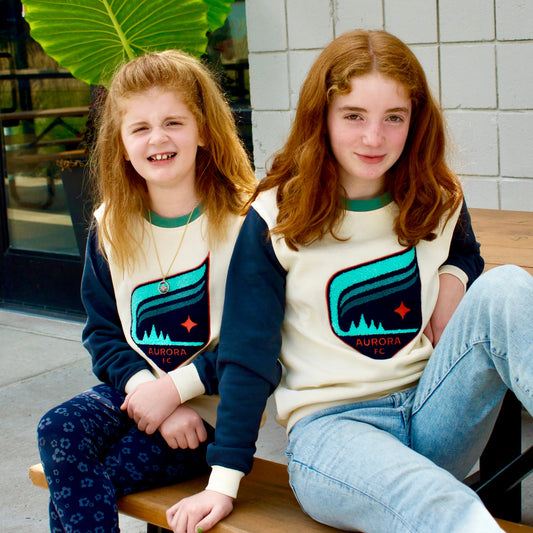 Two young girls sitting on a wooden picnic table outdoors, wearing matching cream and navy long-sleeve shirts with an Aurora Crest logo.