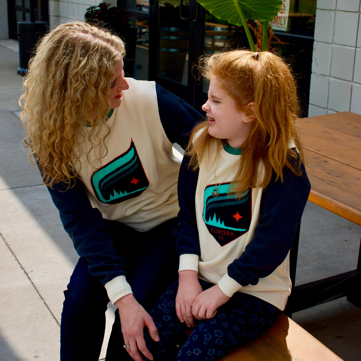 A woman and a young girl sitting on a wooden bench at an outdoor cafe, wearing matching cream and navy long-sleeve sweatshirts with a teal and orange graphic on the front.