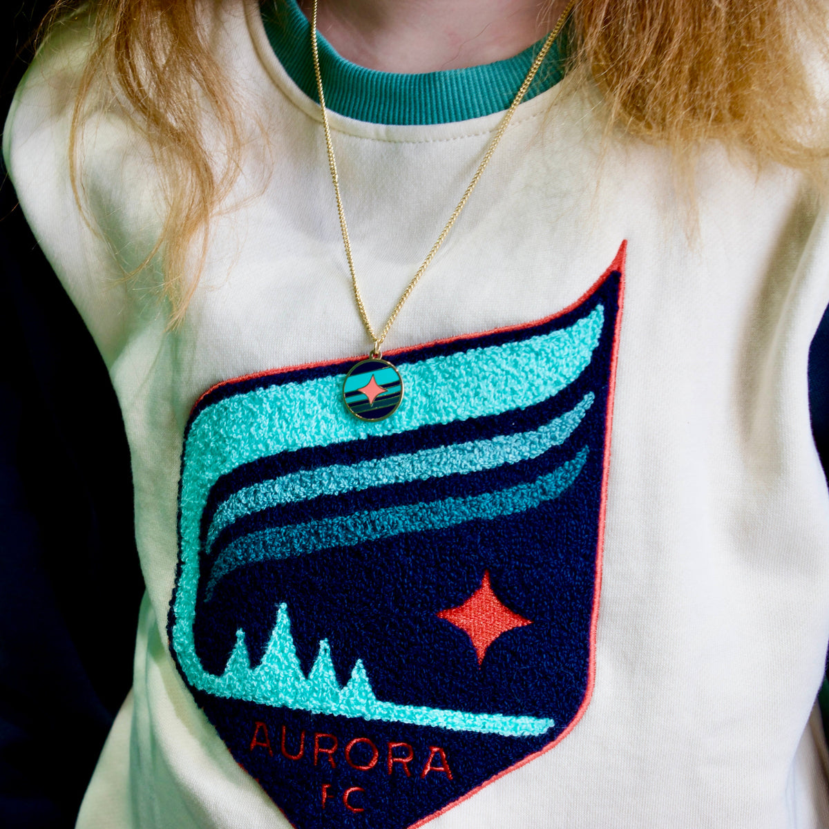A close-up of a person wearing a cream and navy pullover sweatshirt with a large embroidered 'AURORA' graphic on the front. The graphic features turquoise, navy, and red details, and the person is also wearing a gold necklace with a small round pendant.