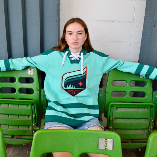 Person wearing a teal hoodie with an aurora crest, sitting among green chairs.