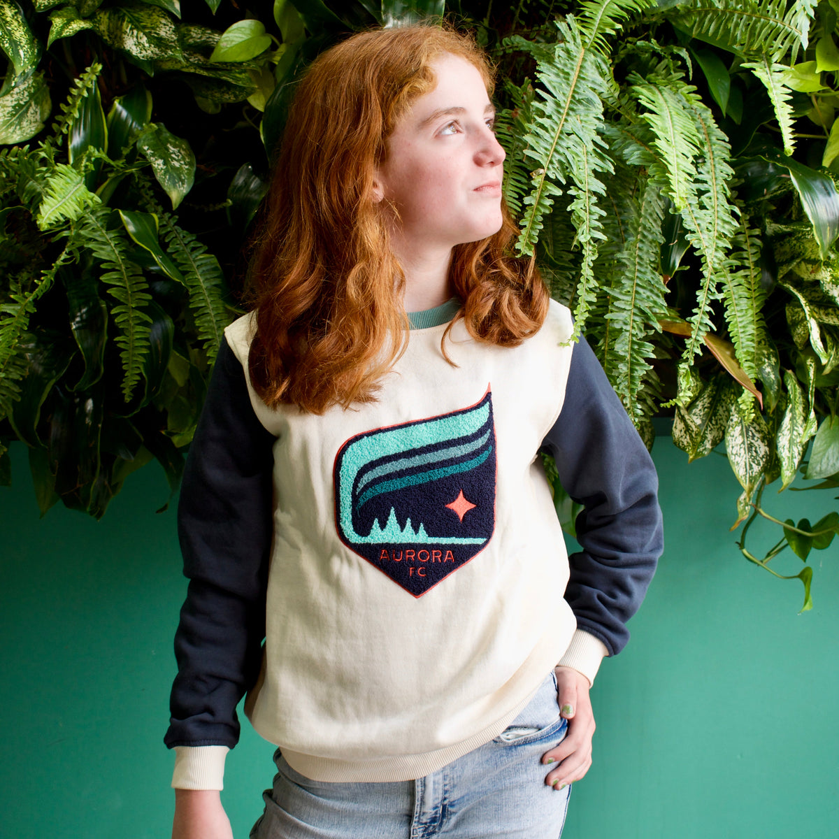 A young woman with red hair wearing a cream and navy long-sleeve sweatshirt with a colorful 'AURORA' patch on the front, paired with light blue jeans, standing in front of a green leaf wall.