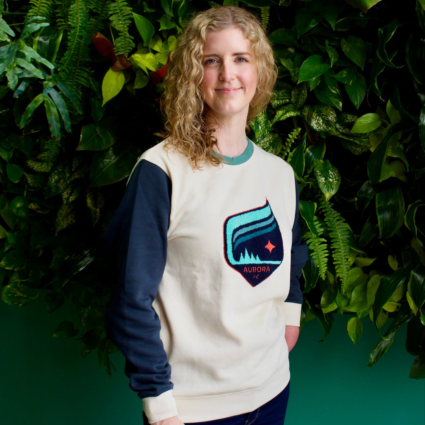 A woman with curly blonde hair stands in front of a green leaf wall, wearing a cream and navy long-sleeve sweatshirt with a colorful shield logo on the chest, paired with dark blue jeans.