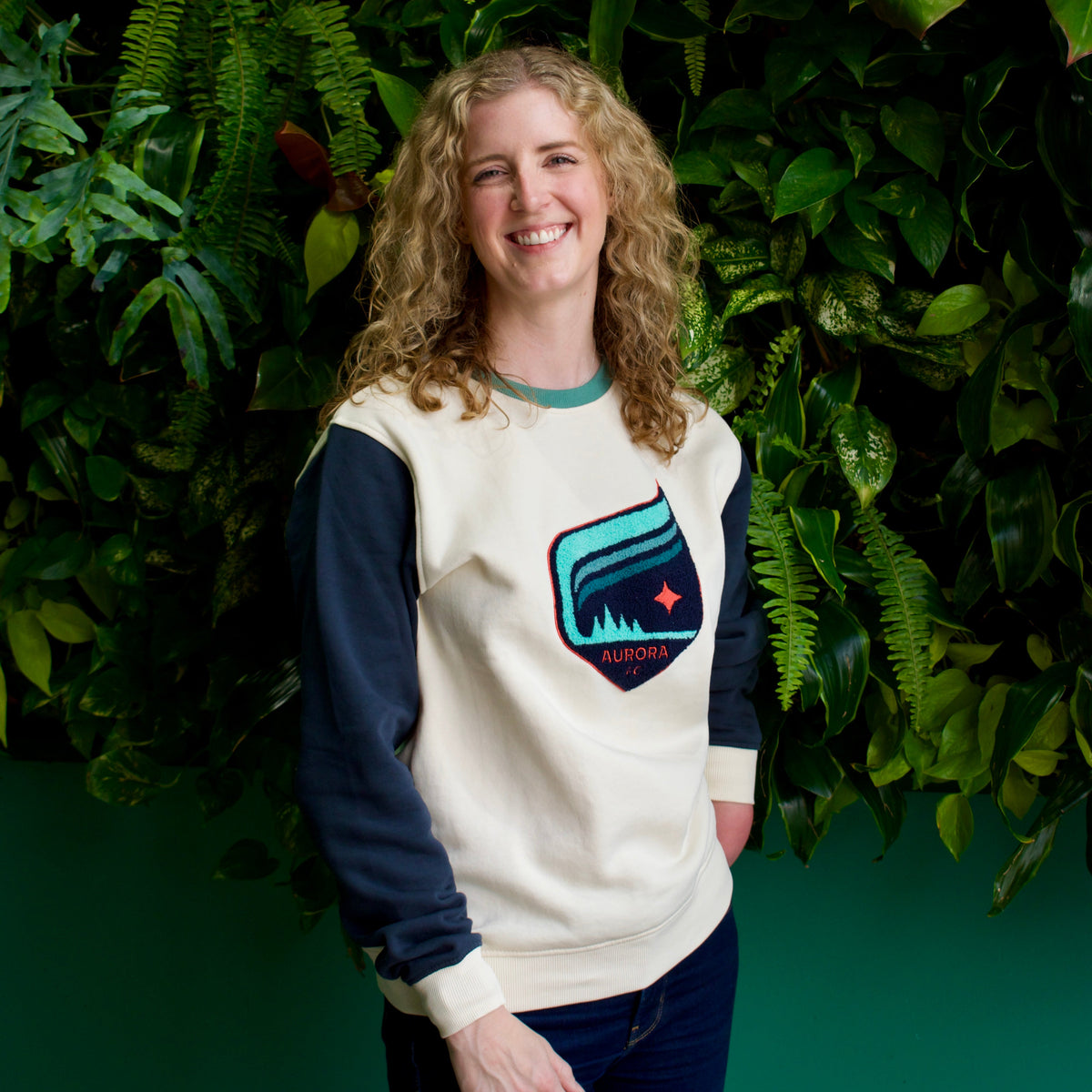 A smiling woman with curly blonde hair stands in front of a green leaf wall, wearing a cream and navy long-sleeve sweatshirt with a colorful shield logo on the chest, paired with dark blue jeans.