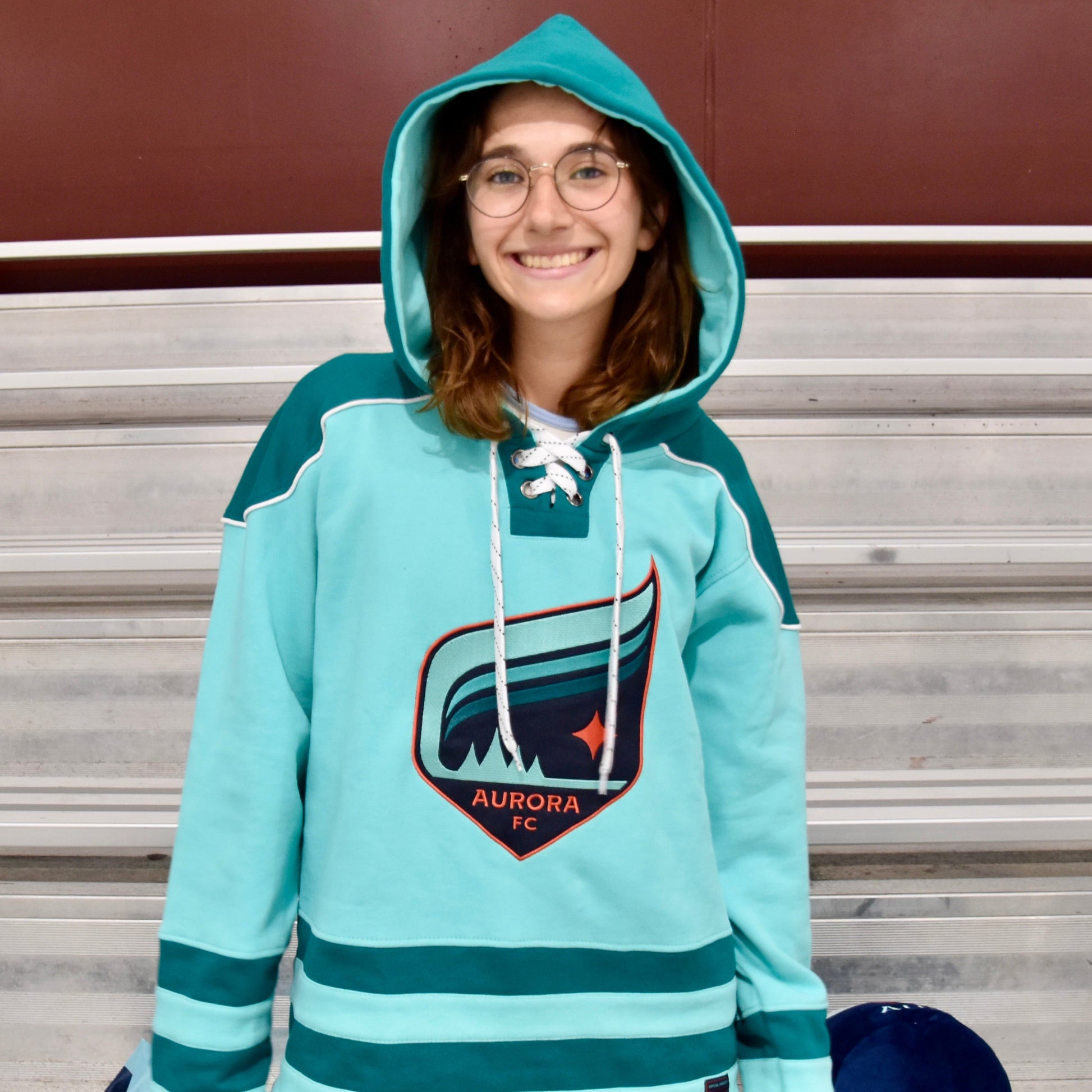 Person wearing a teal and green hoodie with a logo, sitting on bleachers.