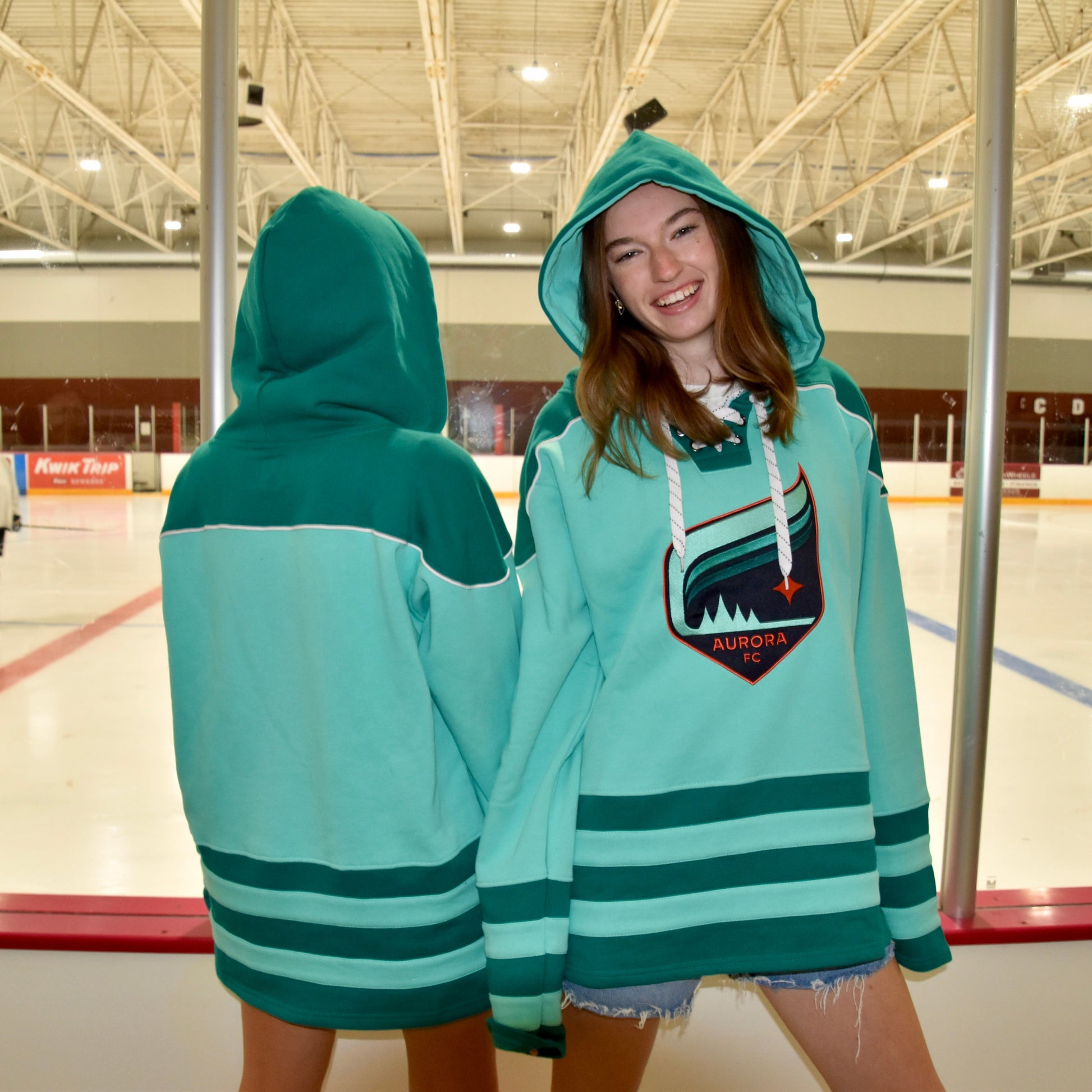 Two people wearing teal hoodies with a logo on an indoor ice rink.