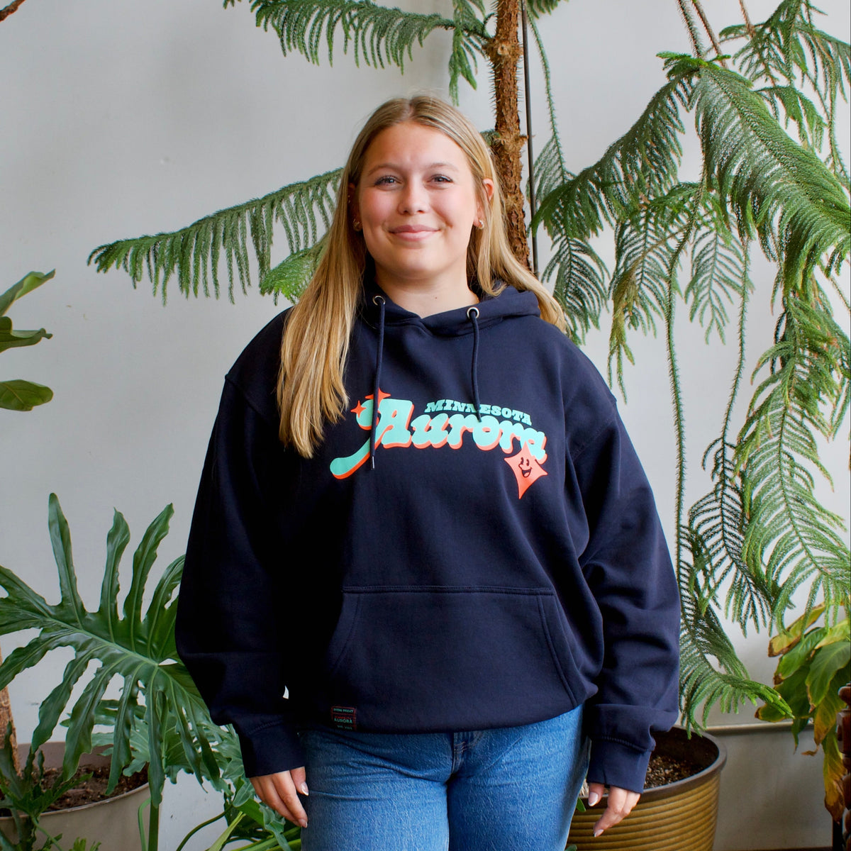 A woman stands indoors wearing a navy blue hoodie with a colorful graphic text print on the chest.The background features large potted green plants against a white wall.