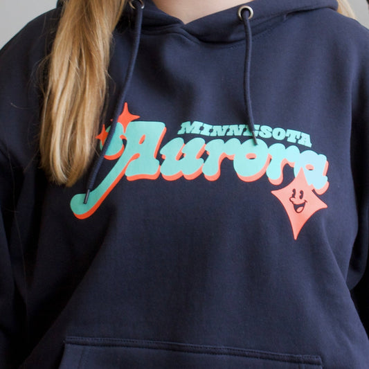 A young woman with blonde hair stands in front of a white wall surrounded by potted green plants, wearing a navy blue hoodie with a colorful 'Flavors' graphic and blue jeans.