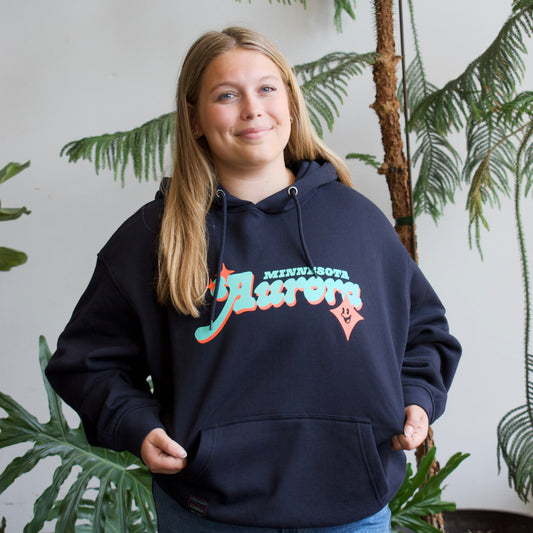 A woman wearing a navy blue hoodie with a colorful 'Aurora' graphic on the chest, standing in front of a white wall with potted plants.