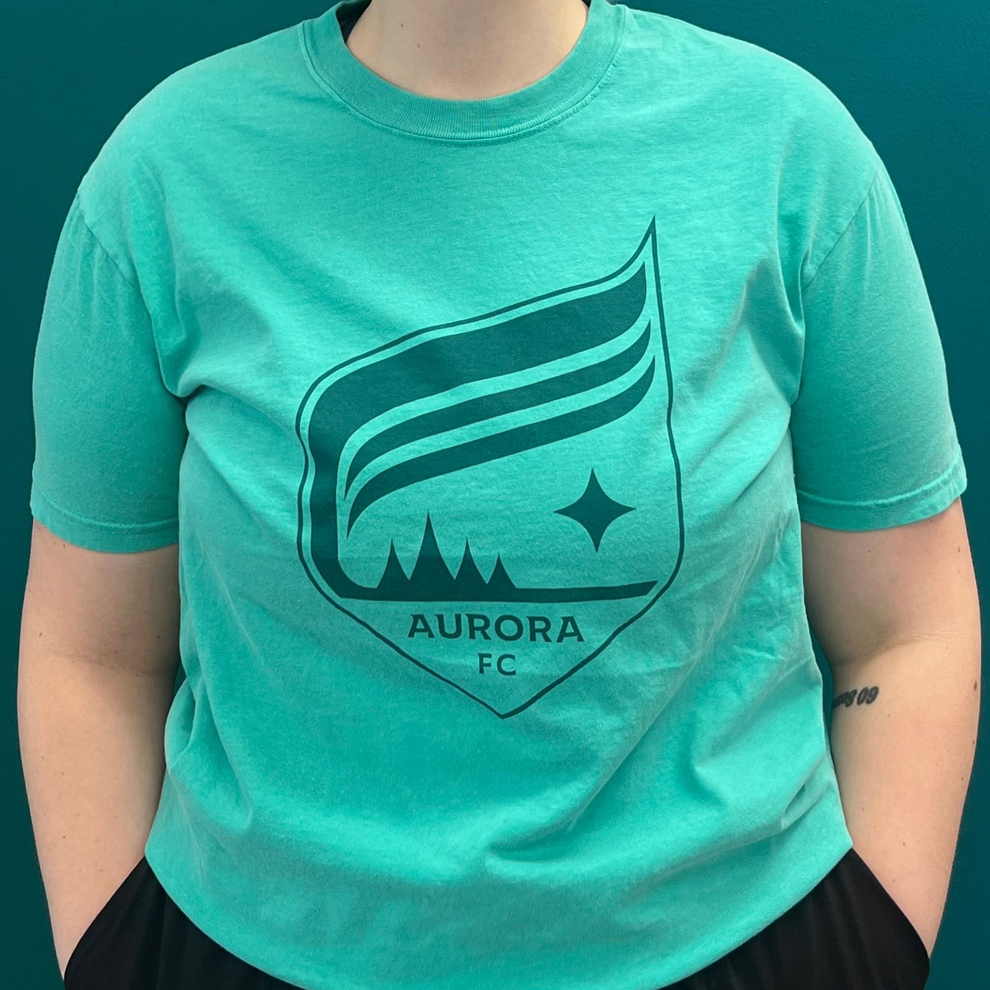 Person wearing a teal t-shirt with 'Aurora FC' logo against a dark green background