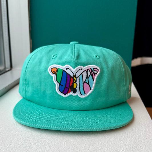 Teal hat with a butterfly that features the words "Queer and Trans" 