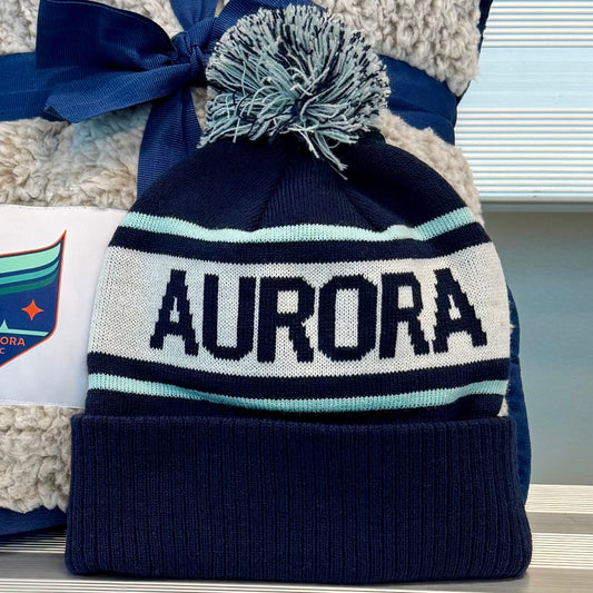 Navy and white beanie with pom-pom and 'Aurora' text, paired with a matching blanket on a metallic surface.