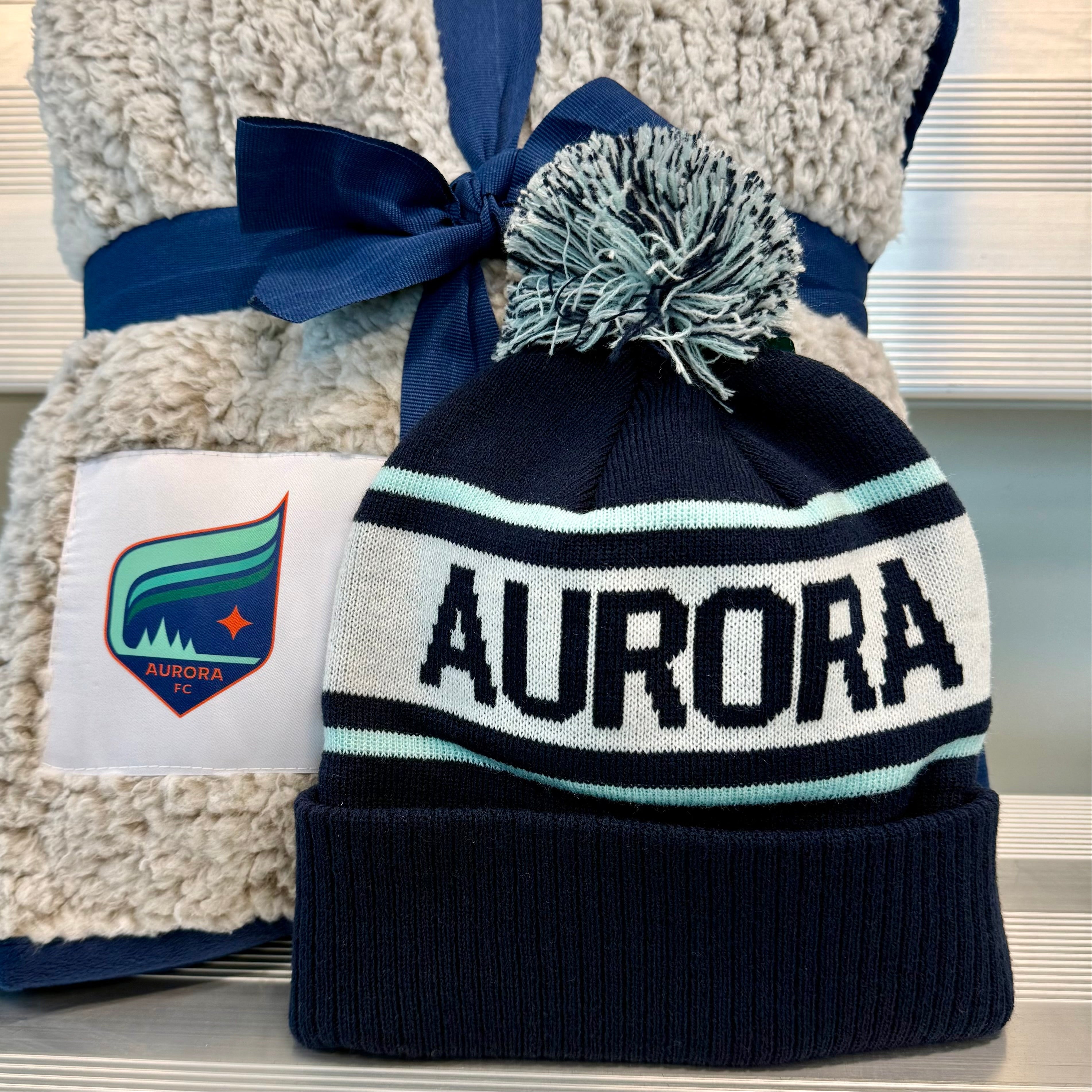 Navy beanie with 'Aurora' text and pom-pom, tied to a plush gray blanket with a visible brand logo.