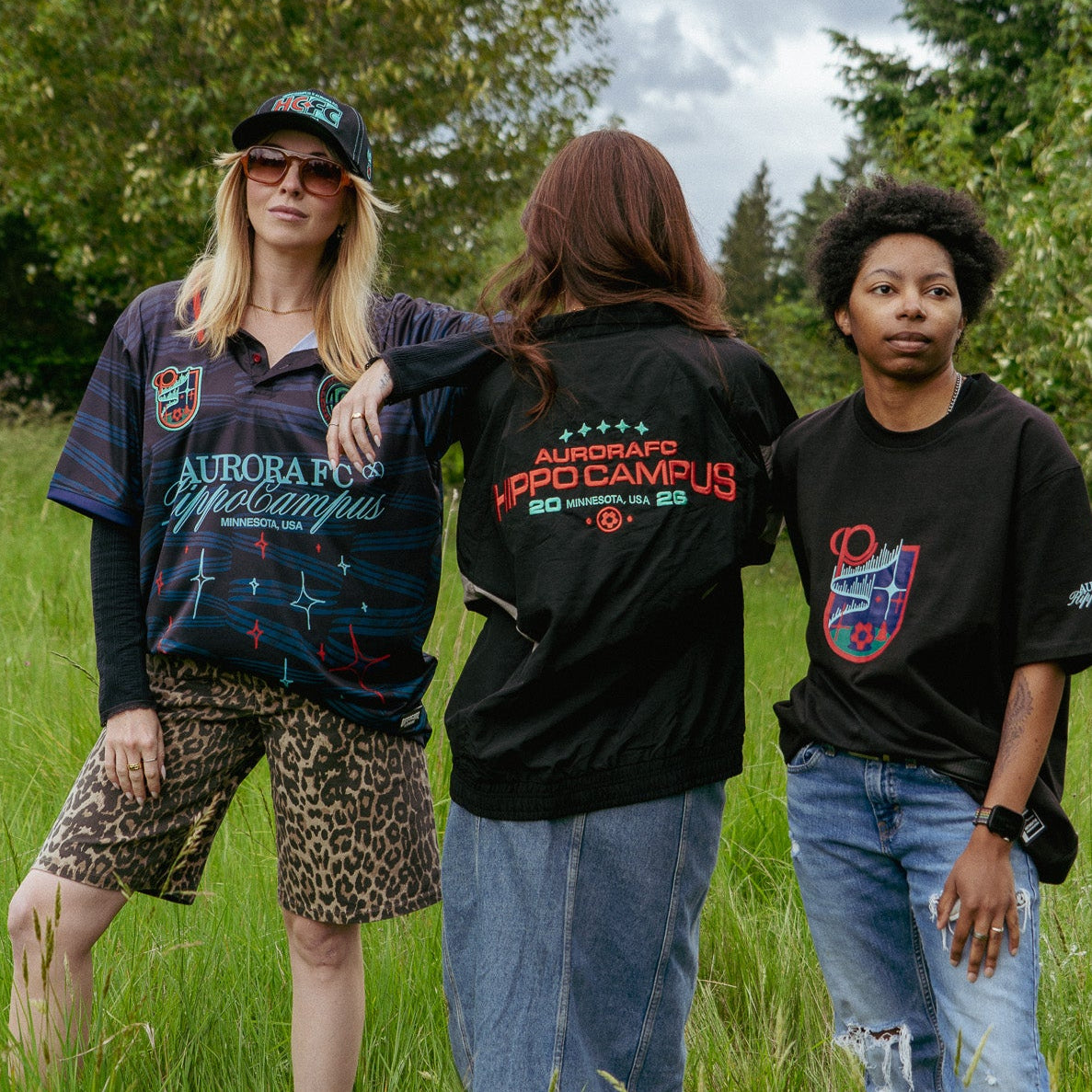 Three people standing in a grassy field wearing black t-shirts with visible text and graphics.