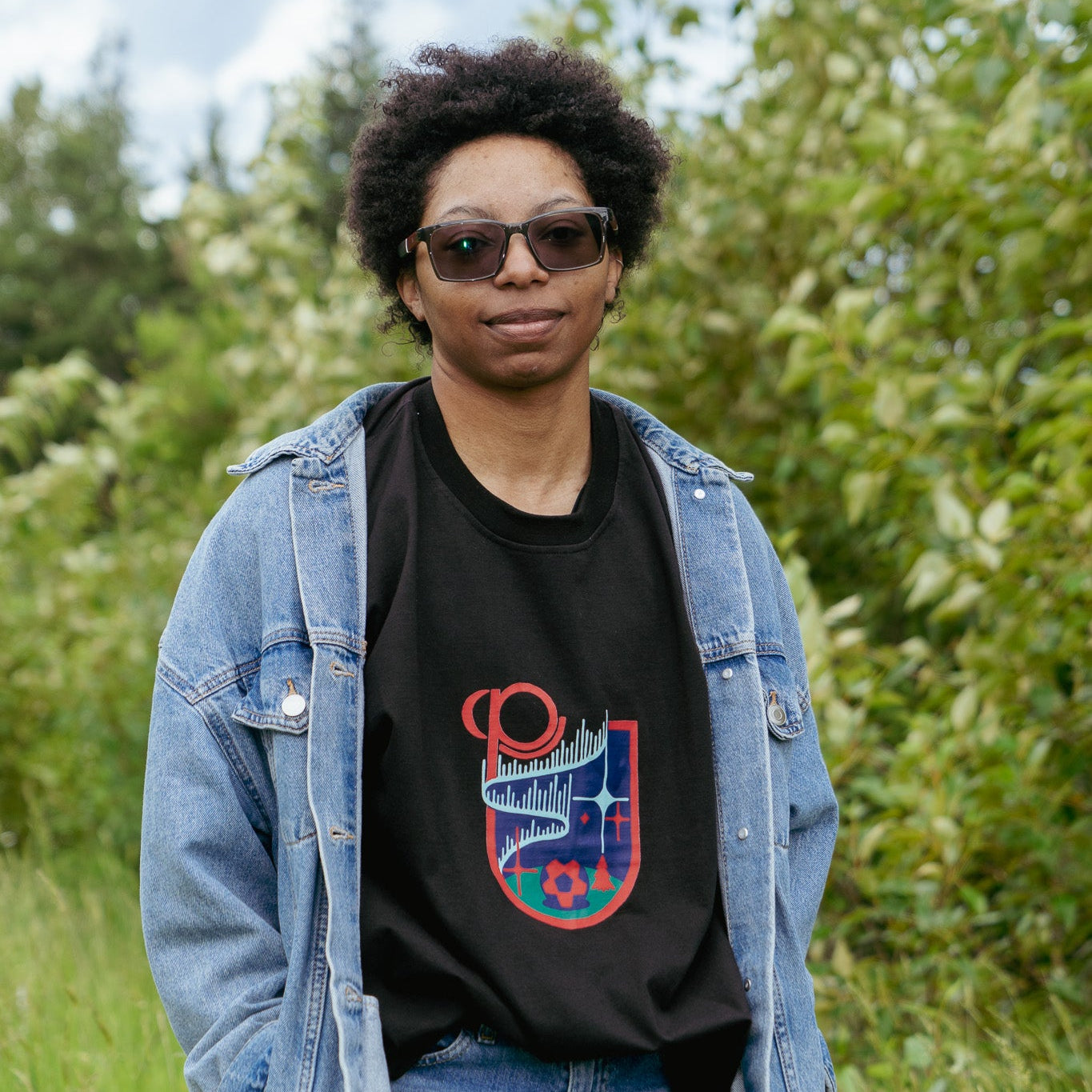 Person wearing a black t-shirt with a colorful design, blue jeans, and a denim jacket outdoors.