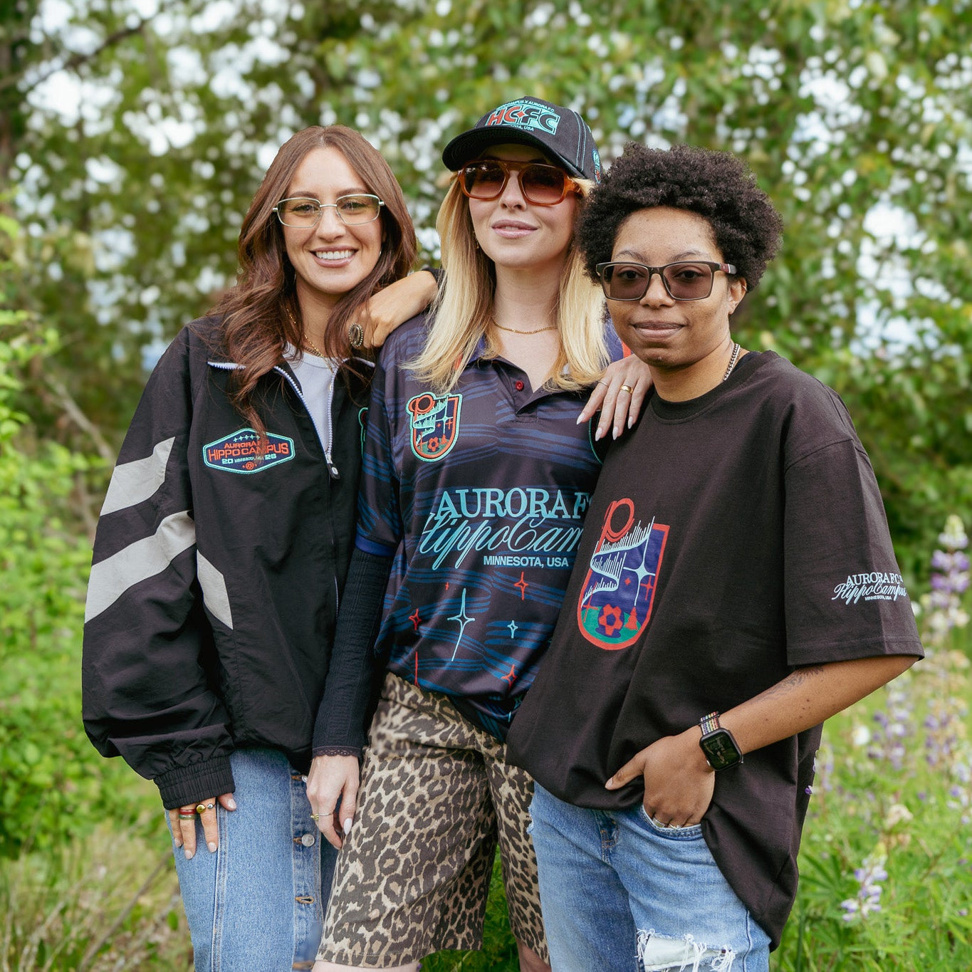 Three women standing together in a field with trees and flowers in the background