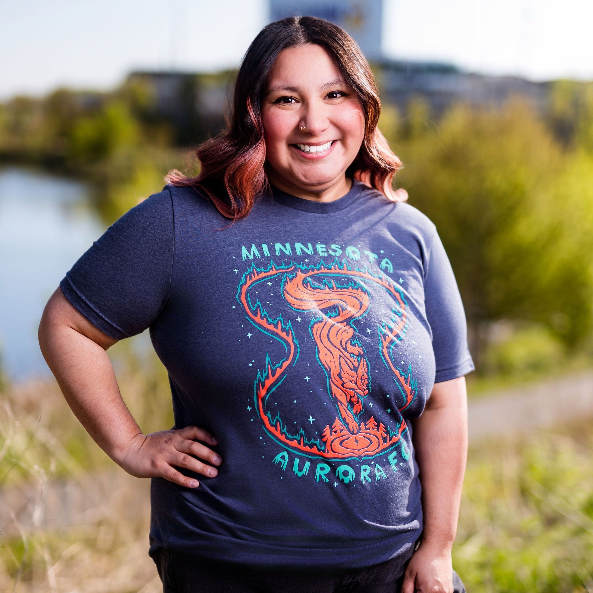 Person wearing a blue t-shirt with a graphic design standing outdoors by a body of water.