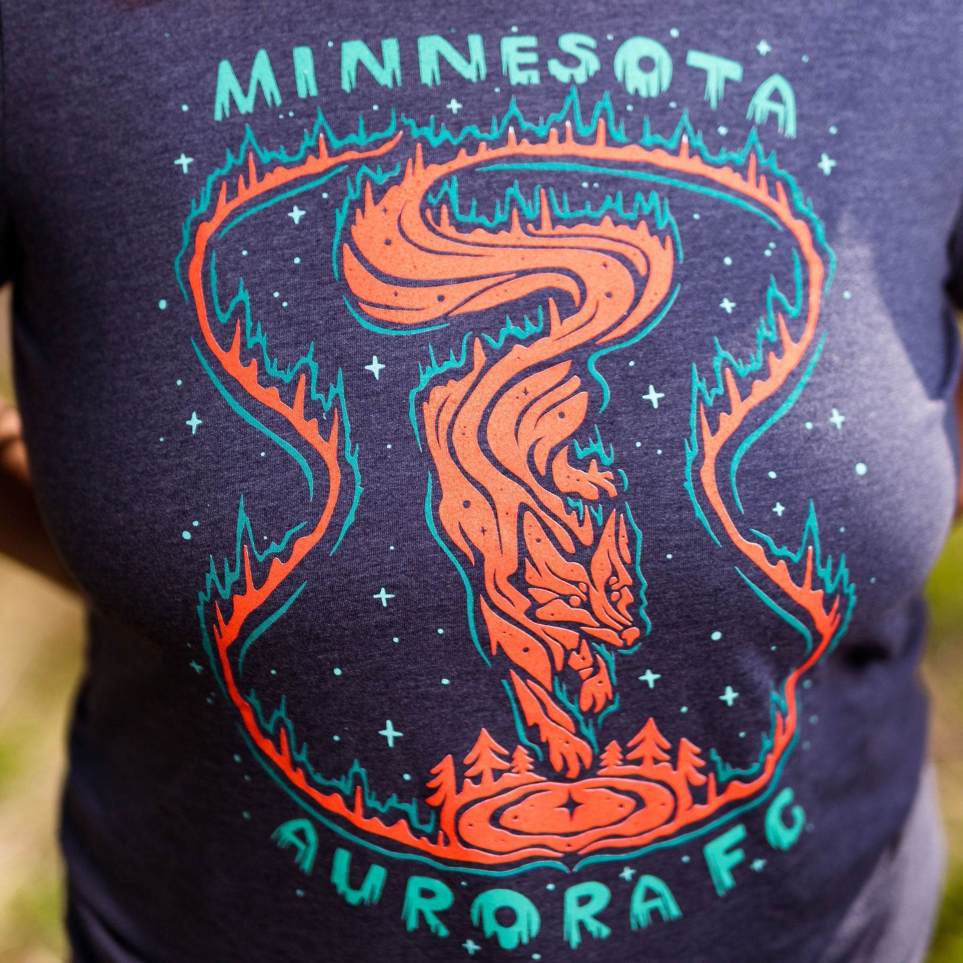 Navy blue t-shirt with orange and teal graphic design featuring 'Minnesota Aurora FC' on a blurred natural background.