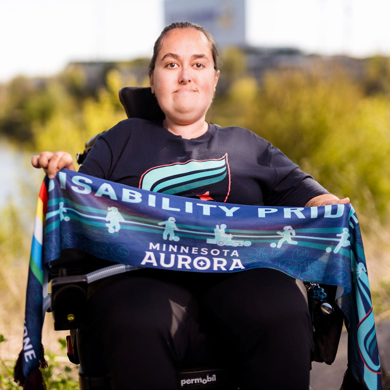 Person in a wheelchair holding a 'Disability Pride' scarf by a lake.