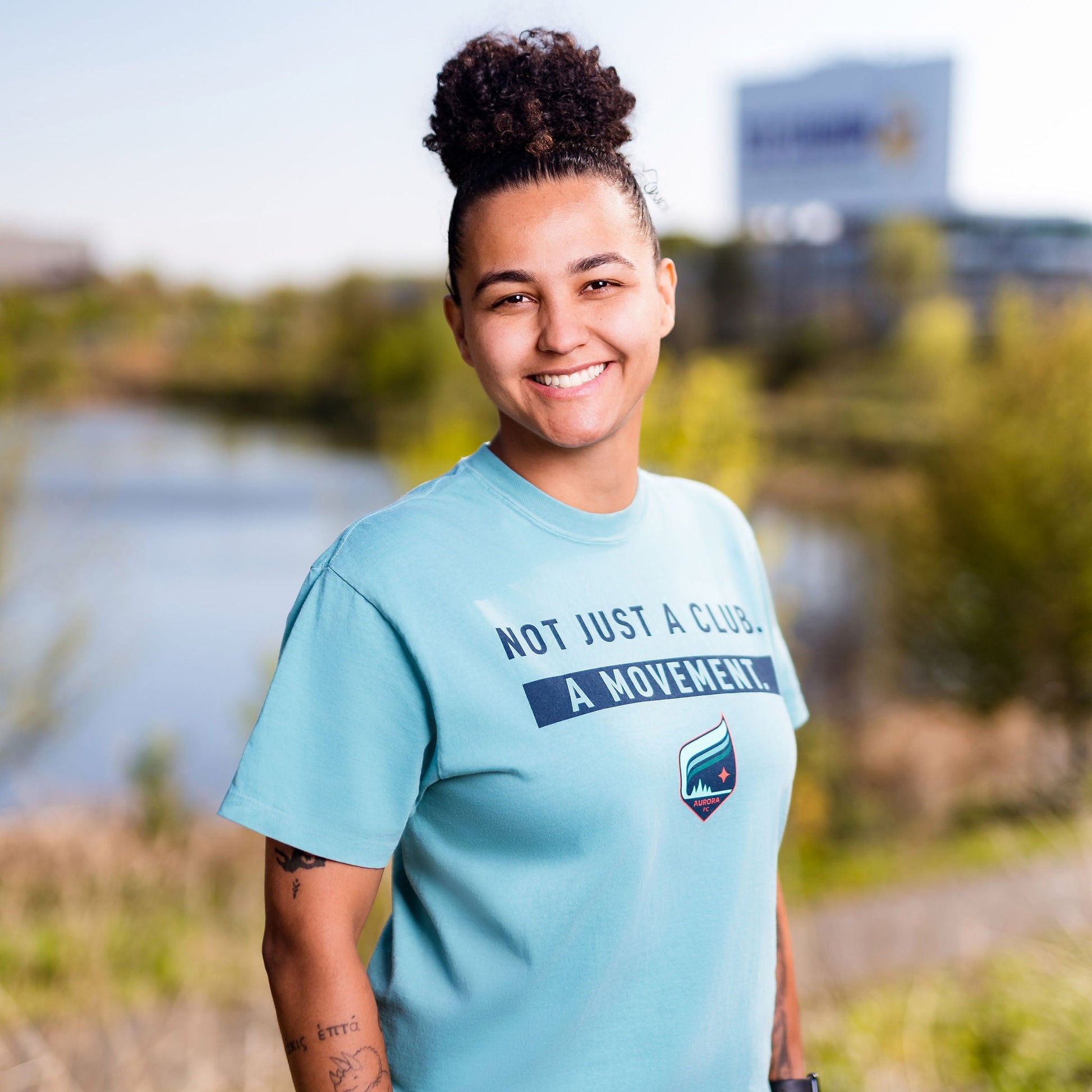 Person wearing a teal t-shirt with text and graphics, standing outdoors near a body of water.